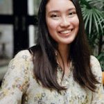 Cheerful young Asian lady with mobile phone in casual clothes smiling and drinking latte while chilling in outdoor cafe
